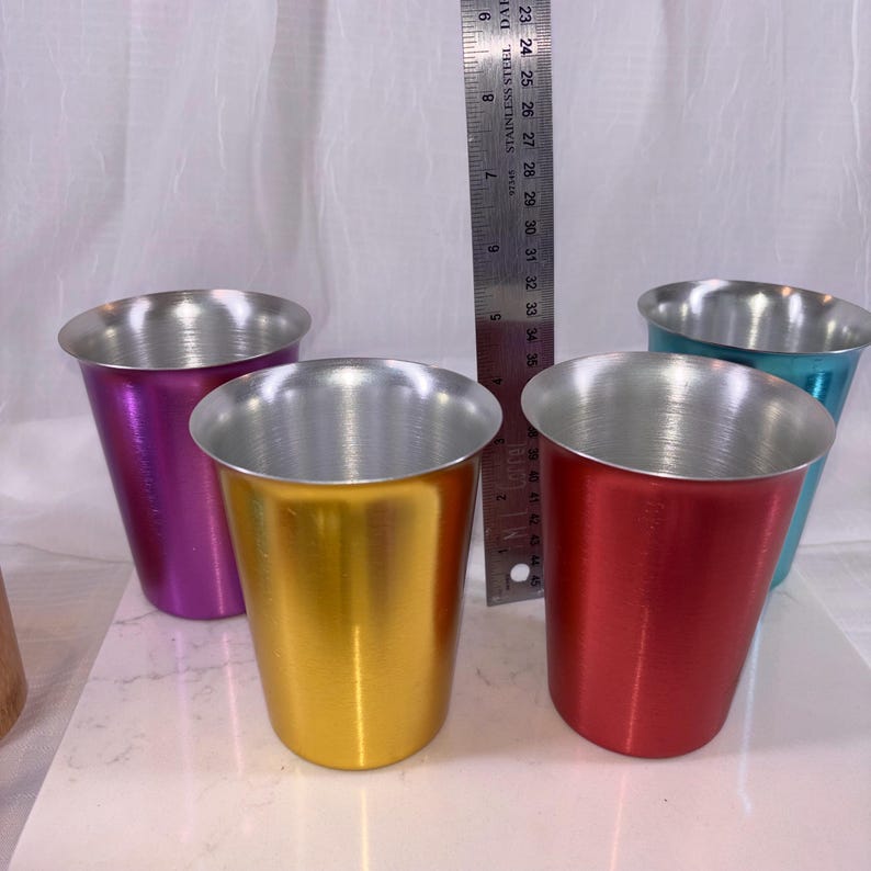 May include: Four colorful metal cups in purple, gold, red, and teal, with a silver interior. A stainless steel ruler is positioned behind the cups. The cups are approximately 3.5 inches tall.