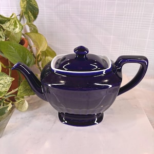 May include: A dark blue ceramic teapot with a white rim and lid. The teapot has a curved spout and handle. The base of the teapot has a textured pattern. A green plant is visible in the background.