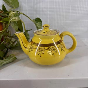 May include: A yellow ceramic teapot with a lid, handle, and spout. The teapot has gold floral and stripe detailing. The teapot is sitting on a white surface. A green plant is in the background.