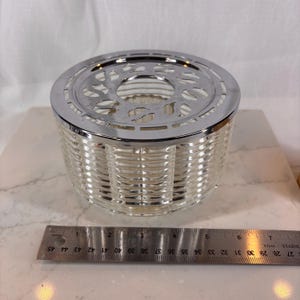May include: A clear glass tea warmer with a woven basket design. It has a shiny silver metal rim and a decorative top with cut-out designs. The warmer is approximately 4 inches in diameter.