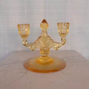May include: A vintage, amber-colored glass candelabra with two candle holders. The candelabra features an ornate, decorative design with a round base and a central, pointed finial. The candle holders are fluted and the piece is made of transparent glass.