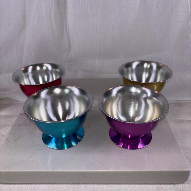 May include: Four small, colorful bowls with a silver interior and a pedestal base. The bowls are in shades of red, gold, teal, and purple. The bowls are sitting on a white surface.