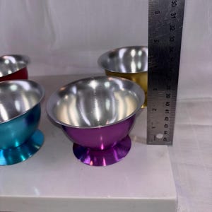 May include: A collection of colorful metal bowls with silver interiors. The bowls are in shades of red, gold, teal, and purple. A ruler shows the bowls are approximately 3 inches tall.