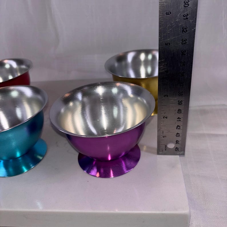 May include: A collection of colorful metal bowls with silver interiors. The bowls are in shades of red, gold, teal, and purple. A ruler shows the bowls are approximately 3 inches tall.