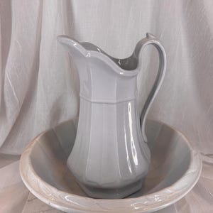 May include: A white ceramic pitcher with a handle and a matching basin. The pitcher has a fluted design and a spout. The basin has a scalloped edge. The set is likely for washing or decorative purposes.