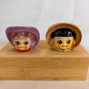 May include: Two ceramic head figurines featuring stylized faces with large blue eyes and rosy cheeks. The left figurine wears a purple hat with curly hair, while the right figurine has black hair and wears a yellow hat with a brown rim.