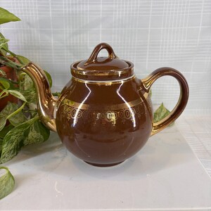 May include: Brown ceramic teapot with a gold-colored handle, spout, and trim. The teapot has a lid and a gold decorative band around the middle. The teapot is sitting on a white surface.