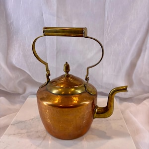 May include: A polished copper tea kettle featuring a hammered texture on the body, a brass lid with a central finial, and a brass handle that arches over the top. The kettle includes a curved brass spout extending from the side of the vessel.