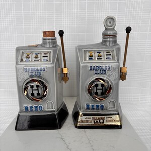 May include: Two gray ceramic decanters shaped like slot machines, with the words "HAROLDS CLUB" and "RENO" in blue. Each has a pull lever and a gold and black emblem. One has a cork stopper, the other a ceramic top.