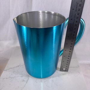 May include: A teal-colored metal pitcher with a handle and a pouring spout. The interior is silver. A ruler is placed next to the pitcher, measuring approximately 8 inches in height. The pitcher sits on a white and gray surface.