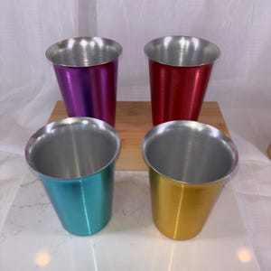 May include: Four colorful metal cups in purple, red, teal, and gold. Each cup has a silver rim and a tapered design. The cups are arranged on a wooden surface.