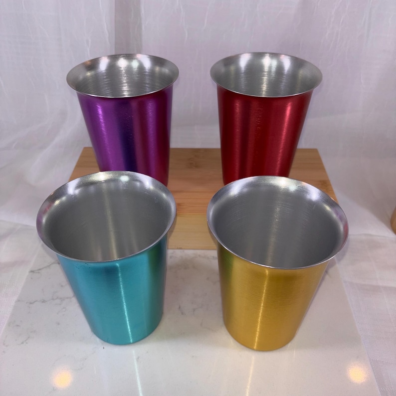 May include: Four colorful metal cups in purple, red, teal, and gold. Each cup has a silver rim and a tapered design. The cups are arranged on a wooden surface.