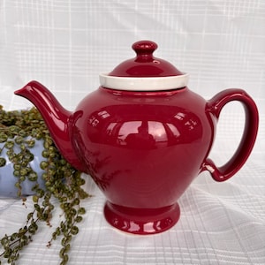 May include: A glossy, burgundy-colored ceramic teapot with a white rim on the lid. The teapot has a curved handle and spout, and sits on a small pedestal base. A small plant is visible in the background.