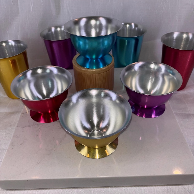 May include: A collection of colorful metal cups and bowls. The bowls have a silver interior and are in various colors, including gold, red, purple, and teal. Some are on pedestals, while others are simple cups.