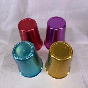 May include: Four metallic tumblers in red, purple, teal, and gold. The cups are arranged in a group, each with a shiny, reflective surface. The tumblers have a classic, tapered shape, suitable for beverages.