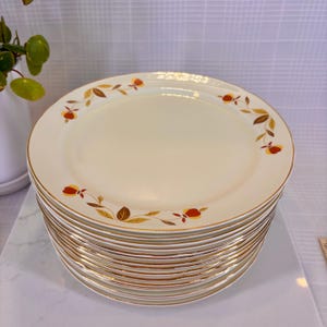 May include: A stack of cream-colored ceramic plates with a gold rim and autumn leaf design. The plates have a slightly oval shape and are stacked on top of each other. The leaf design features red, orange, and brown leaves.