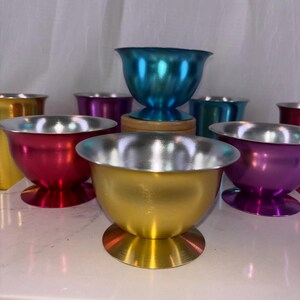 May include: A collection of colorful metal bowls with silver rims. The bowls are in various colors, including red, gold, purple, and teal. Each bowl has a rounded base and a flared rim. The bowls are arranged on a white surface.