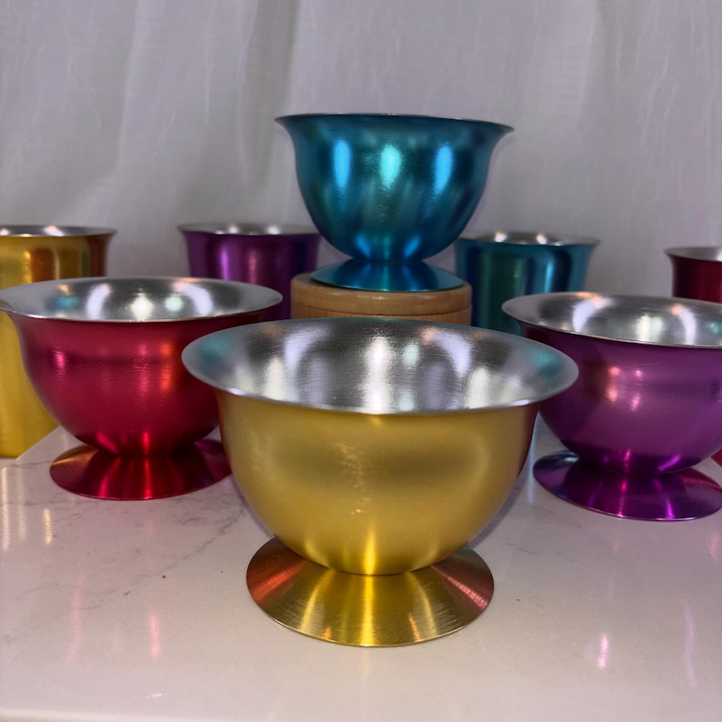 May include: A collection of colorful metal bowls with silver rims. The bowls are in various colors, including red, gold, purple, and teal. Each bowl has a rounded base and a flared rim. The bowls are arranged on a white surface.