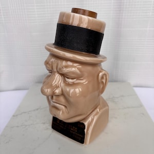 May include: A tan ceramic bust of W.C. Fields wearing a black-banded top hat. The bust has a detailed face with a stern expression. The base has a black label with white text. A wooden piece is on top of the hat.