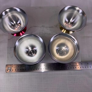 May include: Four small, silver-colored metal bowls with rounded sides and a small base. Two bowls have a red base, and two have a gold base. A ruler is placed below the bowls, with measurements in inches.