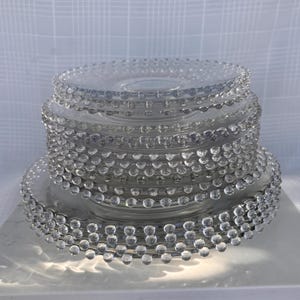 May include: A stack of clear glass plates with a beaded edge design. The plates are round and have a slightly raised center. The glass is transparent, allowing light to pass through, and the beaded edges add a decorative touch.