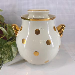 May include: Cream-colored ceramic jar with a lid, adorned with gold polka dots and gold trim. The jar has two gold handles and a matching gold rim on the lid. A decorative piece for the kitchen or dining room.
