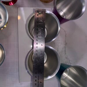 May include: Stainless steel measuring cups, with a ruler showing a measurement of approximately 4 inches. The cups are silver and cylindrical, with a shiny finish. The image is taken from a top-down perspective, showing the interior of the cups.