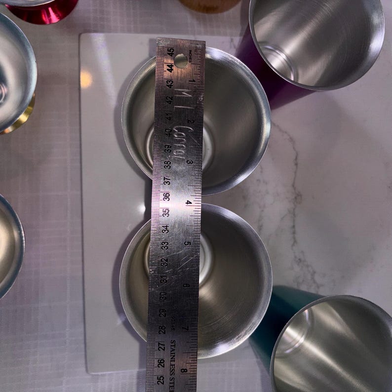 May include: Stainless steel measuring cups, with a ruler showing a measurement of approximately 4 inches. The cups are silver and cylindrical, with a shiny finish. The image is taken from a top-down perspective, showing the interior of the cups.