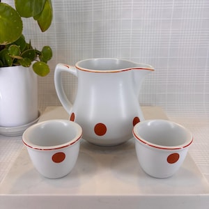 May include: A white ceramic pitcher and two small bowls with red polka dots and red trim. The pitcher has a handle and a spout. The bowls are round and have a similar design. The set is on a white surface.