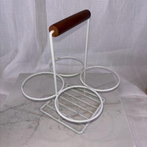 May include: A white metal wine bottle holder with a wooden handle. The holder has four circular openings for bottles and a square base with a grid pattern. The handle is a dark brown color.