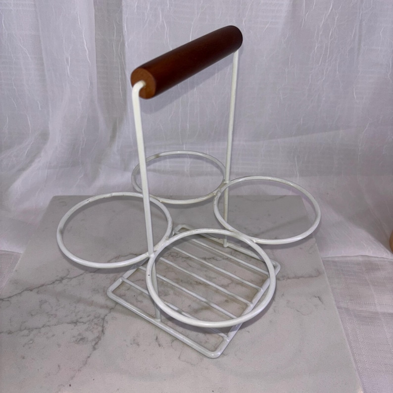 May include: A white metal wine bottle holder with a wooden handle. The holder has four circular openings for bottles and a square base with a grid pattern. The handle is a dark brown color.