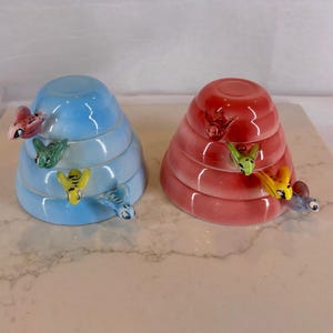 May include: Two ceramic beehive-shaped containers, one light blue and one coral red, each featuring three tiers and small, colorful sculpted bees attached to the exterior surfaces. The glossy finish reflects light on the rounded, stacked forms.