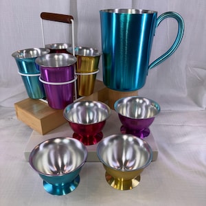 May include: A collection of colorful metal drinkware, including a large teal mug, small footed bowls, and a set of six small cups in a carrier. The drinkware features a shiny, metallic finish in teal, purple, gold, and red. The set is ideal for serving beverages.