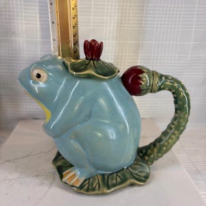 May include: A ceramic teapot shaped like a frog, with a light blue body, yellow mouth, and green and red accents. The handle is a textured green, and the lid is a red flower. The teapot sits on a green lily pad base.