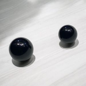 May include: Two black spheres of different sizes, both with a glossy finish, are displayed on a white surface.