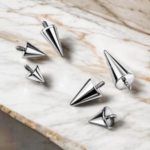 May include: Five silver-toned, pointed, cone-shaped body jewelry pieces. The jewelry pieces are of varying sizes and are arranged on a marble surface.