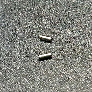 May include: Two small, silver-colored screws with threaded shafts are positioned on a textured, dark gray surface. The screws are cylindrical and appear to be made of metal. The image is a close-up shot.