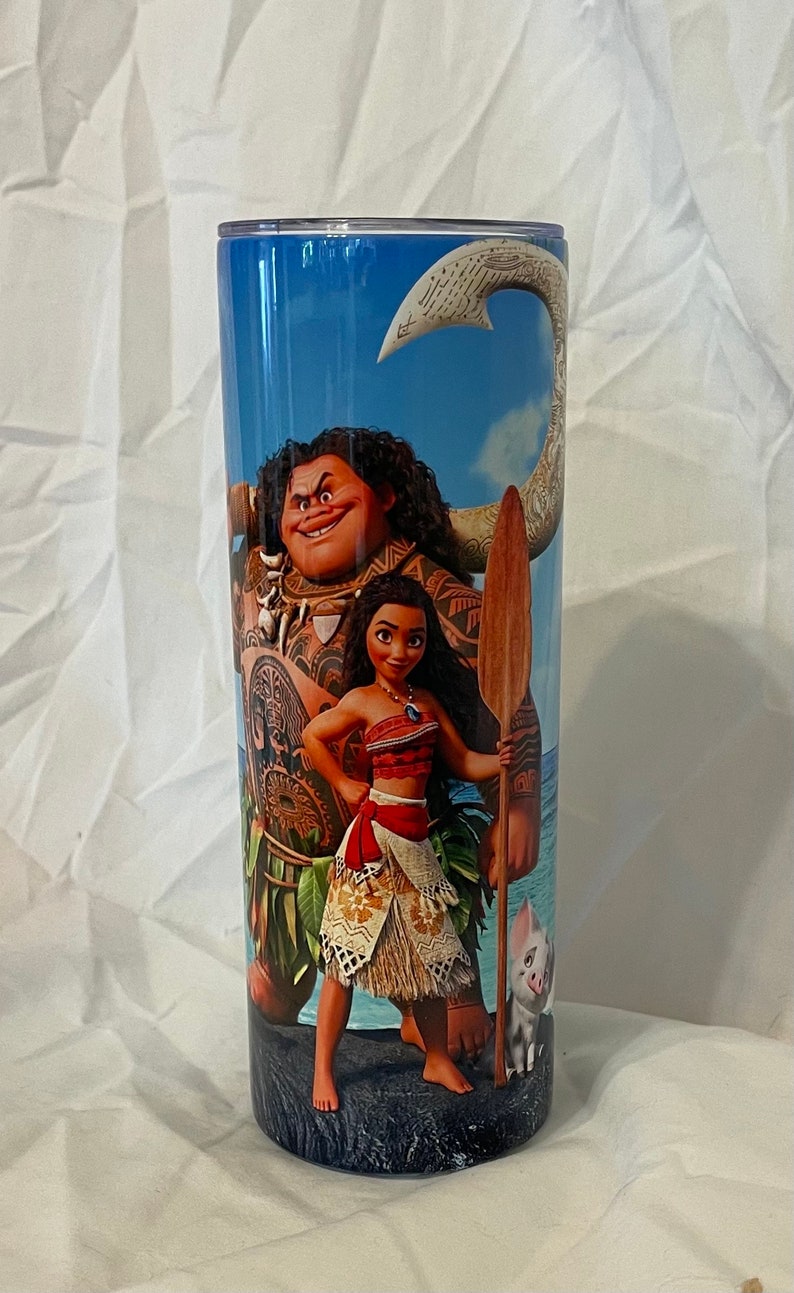 K&ouml;nnte beinhalten: Ein Edelstahlbecher mit einem farbenfrohen Bild von Moana und Maui aus dem Disney-Film Moana. Das Bild zeigt Moana, die vor Maui steht, der eine Lanze h&auml;lt. Ein kleines Schwein steht hinter Moana.