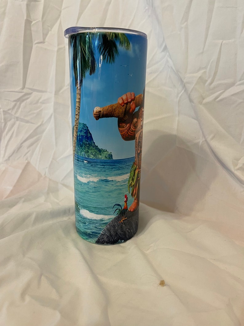 K&ouml;nnte beinhalten: Ein hoher, rostfreier Becher mit einem bunten Bild eines Zeichentrickcharakters aus dem Film Vaiana. Das Bild zeigt eine tropische Inselszene mit Palmen, Ozeanwellen und einem Hahn.