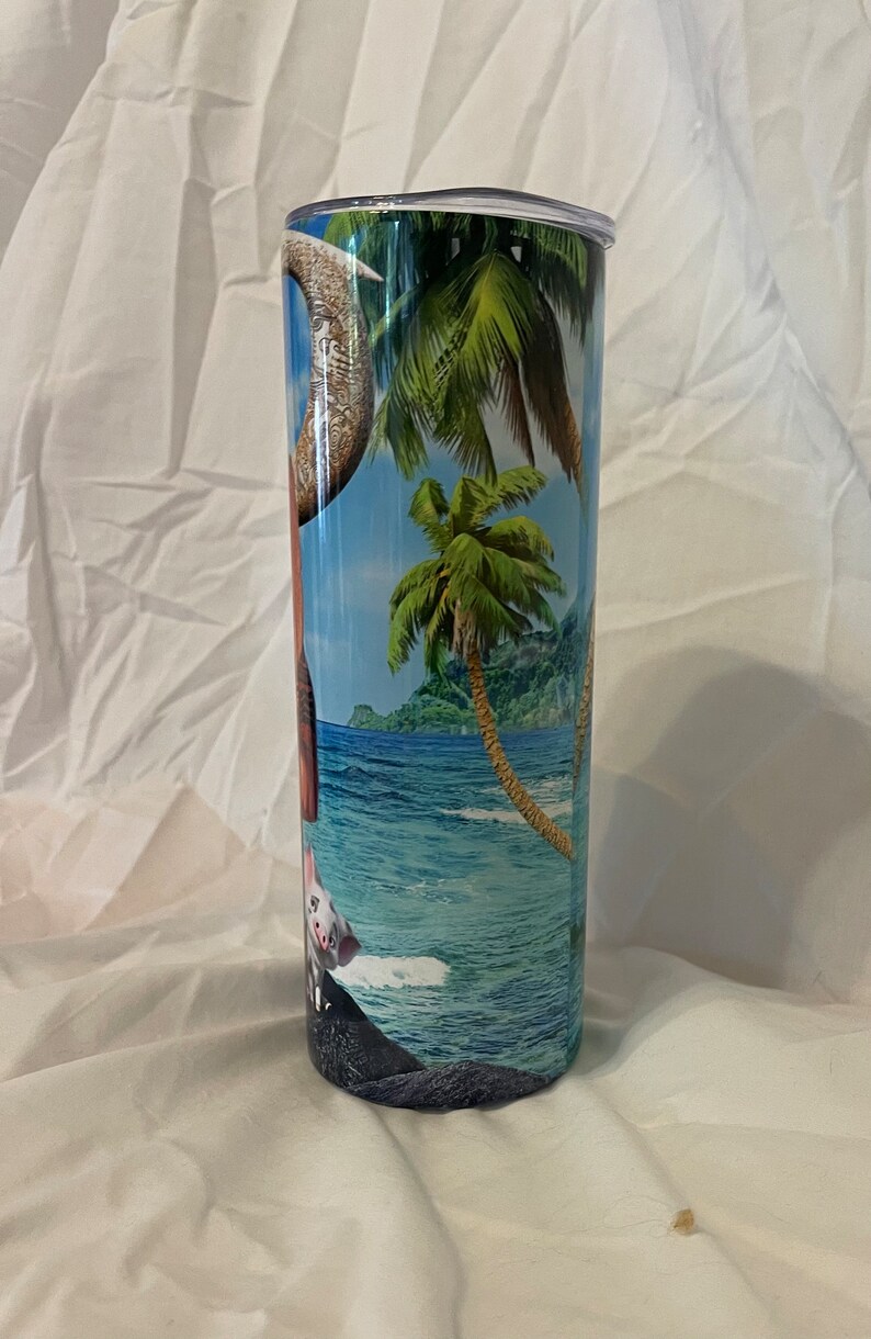 K&ouml;nnte beinhalten: Ein Edelstahlbecher mit einer tropischen Strandkulisse mit Palmen, blauem Wasser und einem Schwein. Der Becher hat einen durchsichtigen Deckel.