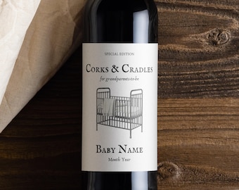 Può includere: Una bottiglia di vino scuro con un'etichetta che recita "Corks & Cradles" e "Baby Name". L'etichetta presenta un disegno di una culla. La bottiglia è su una superficie di legno con un panno bianco.