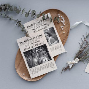 以下が含まれることがあります： 「The Bridesmaid Times」の見出しと「Will You Be My Bridesmaid?」と「Will You Be My Maid of Honor?」の質問が書かれた新聞スタイルのカード2枚が、木製のトレイの上に置かれ、緑と真珠が添えられています。