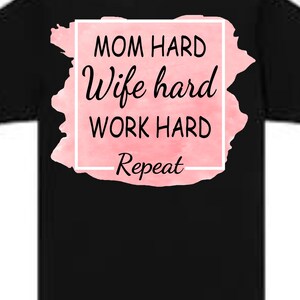 May include: Black t-shirt with a pink watercolor design. The design features the words "MOM HARD, Wife hard, WORK HARD, Repeat" in a white box with black text. A casual tee for everyday wear.