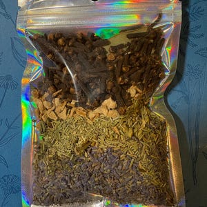 Puede incluir: Una bolsa transparente e iridiscente sellada, llena de varias hierbas y especias secas. La bolsa contiene clavos de olor marrón oscuro, jengibre marrón claro, romero verde y lavanda púrpura. La bolsa está sellada con un cierre de cremallera y tiene un pequeño agujero en la parte superior.