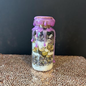 May include: A small glass bottle filled with dried herbs, flowers, and crystals. The bottle is sealed with a cork and purple wax. The wax drips down the sides of the bottle. The contents are visible through the clear glass.