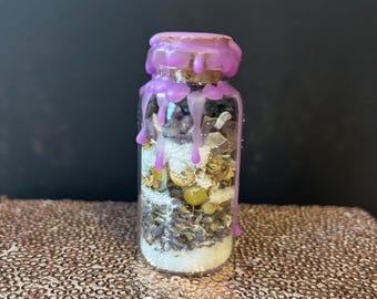 May include: A small glass bottle filled with dried herbs, flowers, and crystals. The bottle is sealed with a cork and purple wax. The wax drips down the sides of the bottle. The contents are visible through the clear glass.