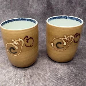 May include: Two ceramic cups with a light brown exterior and a white interior. Each cup features a decorative design in cream and dark red. The rim of each cup has a blue and white geometric pattern.