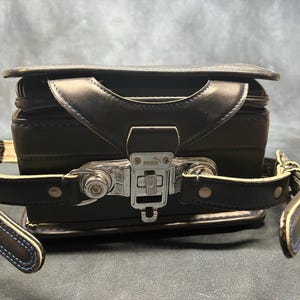 May include: Black leather school bag with a silver clasp and handle. The bag has a rectangular shape with a flap closure and adjustable straps. The straps have white stitching and blue trim.