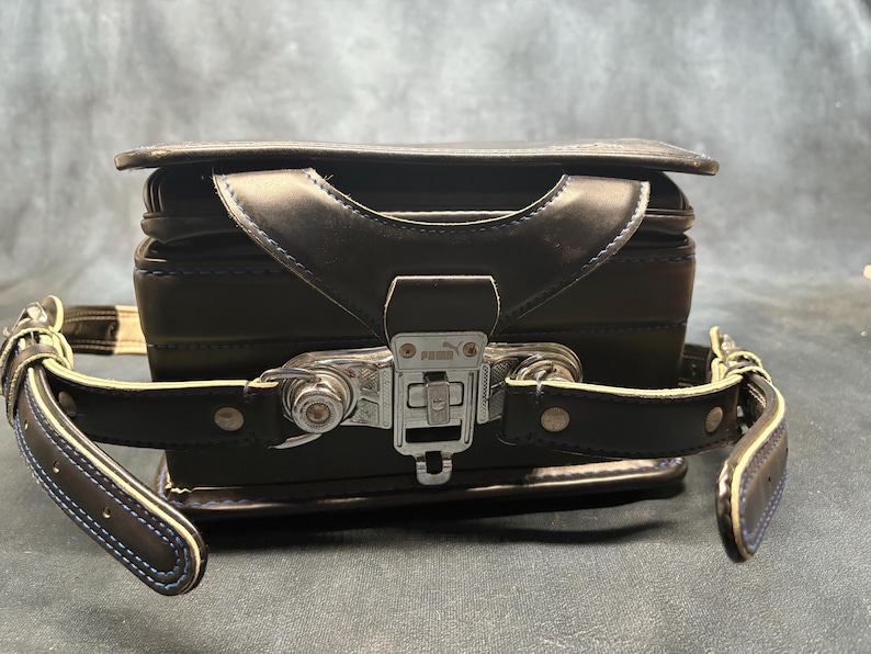 May include: Black leather school bag with a silver clasp and handle. The bag has a rectangular shape with a flap closure and adjustable straps. The straps have white stitching and blue trim.