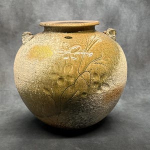 May include: A round, handcrafted ceramic vase with a textured, earthy appearance. The vase features a light brown and tan colour palette with embossed floral designs and small handles. The vase is approximately 25 cm in diameter.